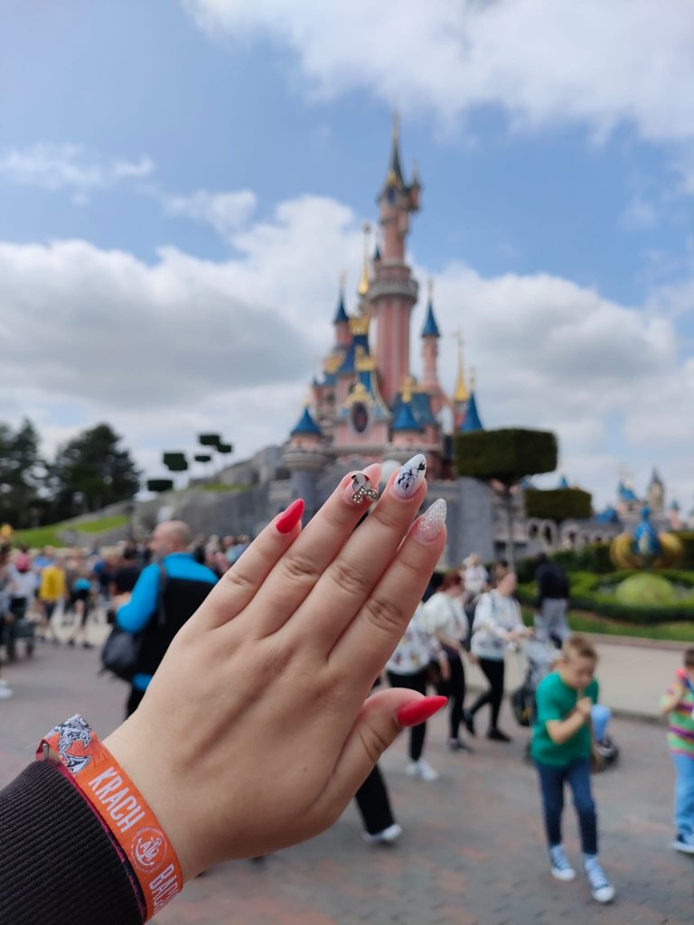 Blick auf das Disney-Nageldesign. Die Hand wird frontal in die Kamera gehalten. Im Hintergrund ist das Dornröschen-Schloss im Disneyland Paris zu sehen.